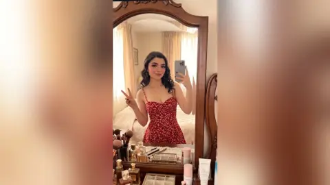 Young woman in a red floral sundress taking a selfie in a bedroom mirror with a cheerful mood.