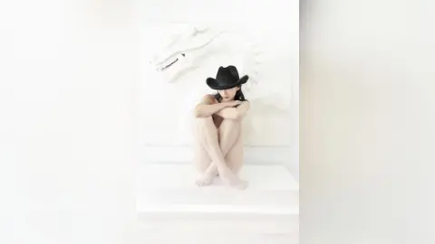 Fashionable woman in a black cowboy hat sitting cross-legged against a white minimalist wall background.