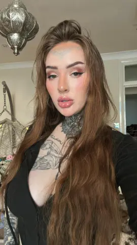 Young woman with long brown hair and neck tattoos wearing a black top in a cozy room with bohemian decor.