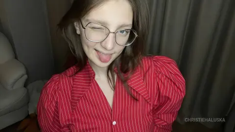 Playful young woman with glasses wearing a red pinstripe blouse in a cozy indoor setting.