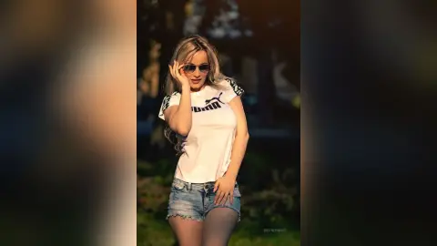 Stylish woman in a white Puma t-shirt and denim shorts posing outdoors in warm golden hour sunlight.