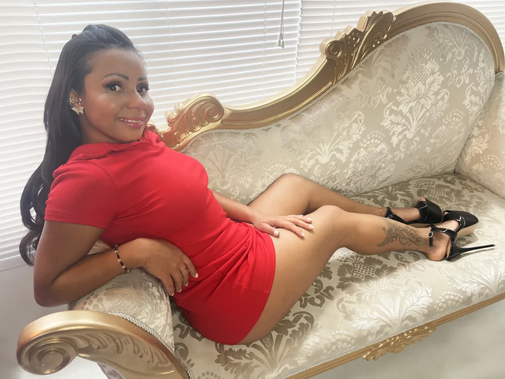 Alondra_Montaner — Alondra_Montaner smiles while reclining on a vintage gold and cream sofa in a tight red mini dress.