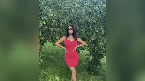 Woman in a red sleeveless dress posing in a lush green apple orchard during a sunny summer day.
