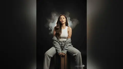 Young woman in white tank top and denim jeans sitting against a moody dark background with ethereal smoke.