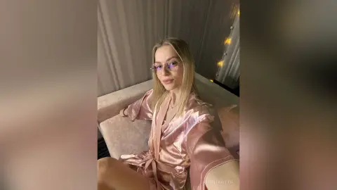 Blonde woman in a pink satin robe relaxing on a bed in a cozy, dimly lit bedroom setting.