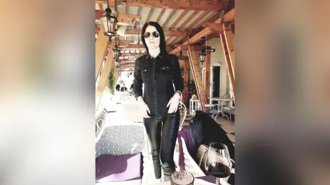 DouceLeya wears black sunglasses, a black button-up shirt, and tight black leather pants while standing outdoors under a wooden patio roof.
