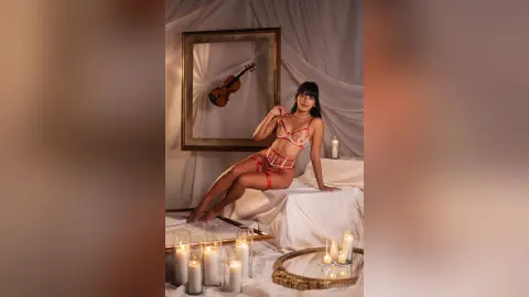 Elegant woman in red lace lingerie sitting on white fabric near glowing candles in a moody, romantic setting.