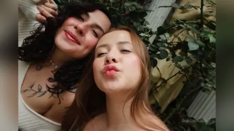 Two smiling women in white tops posing closely together in a lush green garden setting.