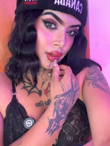 Woman with wavy black hair and tattoos wearing a black lace camisole and Canada beanie in soft pink lighting.