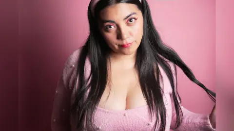 Young woman with long dark hair wearing a pink fuzzy sweater against a soft pink background.
