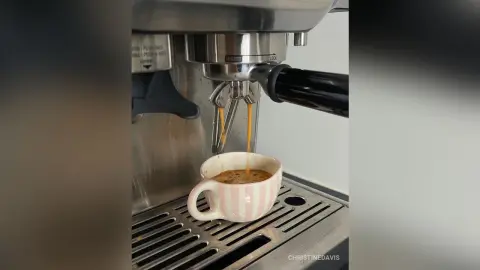 ChristineDavis prepares coffee using a silver espresso machine, pouring a stream of espresso into a pink and white striped mug.