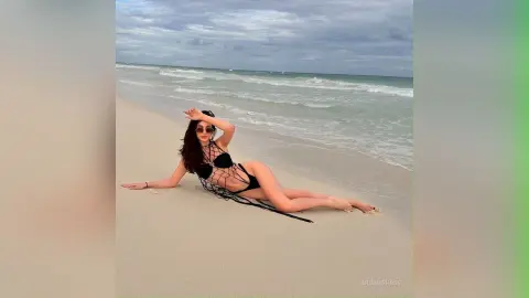 Woman in black bikini and mesh cover-up relaxing on a sandy beach under a cloudy sky.