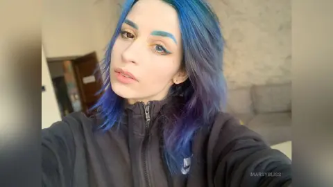 Young woman with blue hair and winged eyeliner wearing a black Nike sweatshirt in a cozy indoor setting.