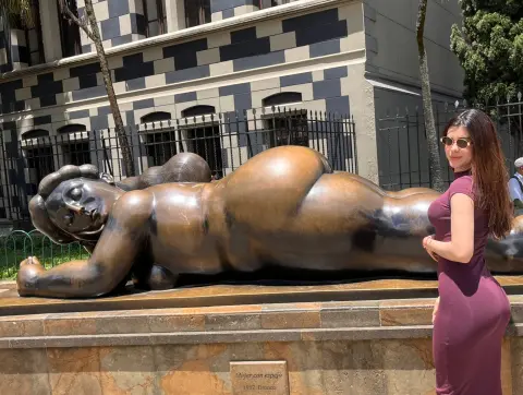 Woman in a purple dress standing next to a large bronze reclining female sculpture in a sunny outdoor plaza.