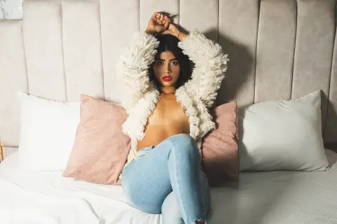 Young woman in a white faux fur jacket and blue jeans sitting on a bed with pillows in a cozy bedroom setting.