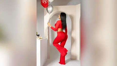 Woman in a red long-sleeve crop top and matching trousers holding birthday balloons in a minimalist white room.