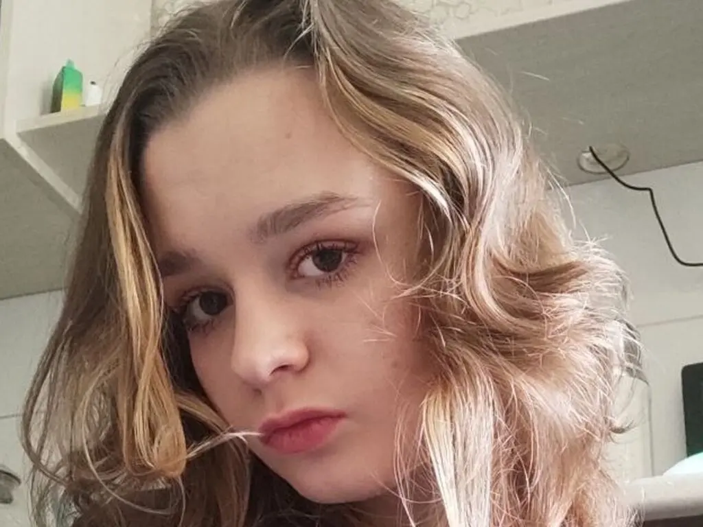 Close-up portrait of a young woman with wavy blonde hair looking thoughtfully at the camera in a bright room.