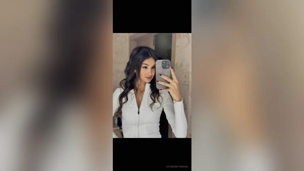 CatherineThomase — CatherineThomase takes a mirror selfie wearing a tight white long-sleeve top and long wavy dark hair.