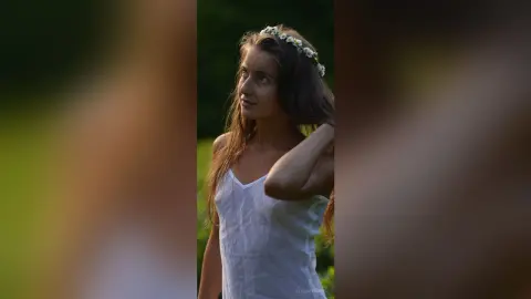 Dreamy portrait of a young woman in a white summer dress and flower crown in a lush green field.