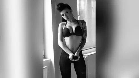 Moody black and white portrait of a tattooed woman in a black bra and leggings standing by a bright window.