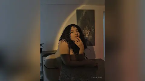 EmilyJune poses in a dimly lit room, leaning over a counter with her long curly hair falling over her shoulders.