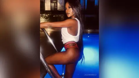 Stylish woman in a white tank top and red shorts posing by a glowing blue swimming pool at night.