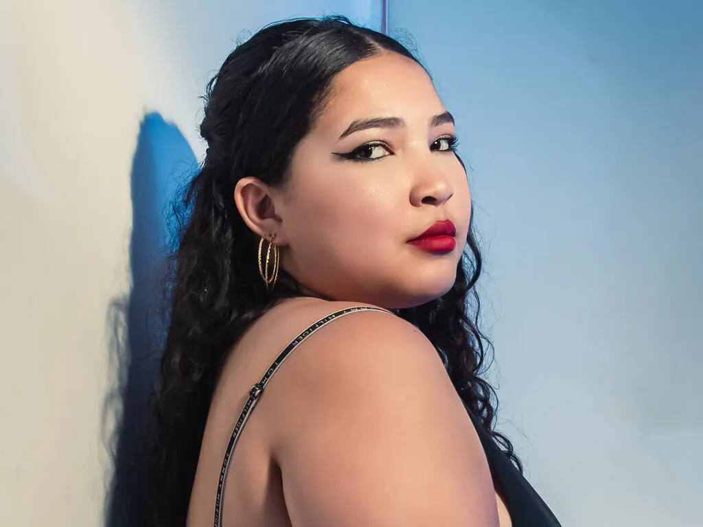 SusanVali poses with her long dark hair and red lipstick, wearing gold hoop earrings and a black top against a plain blue wall.