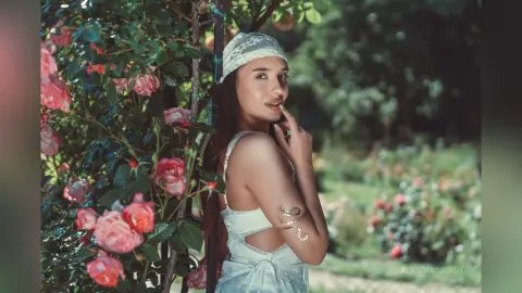 AlessiaCrush poses in a white lace outfit while leaning against a rose bush in a garden.