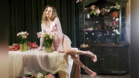 SashaLune poses sitting on a table in a pink dress and thigh-high stockings, surrounded by pink and white flowers in a dark room.