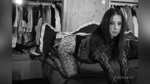 Moody black and white portrait of a woman in patterned lace bodysuit lounging on a bed in a bedroom setting.