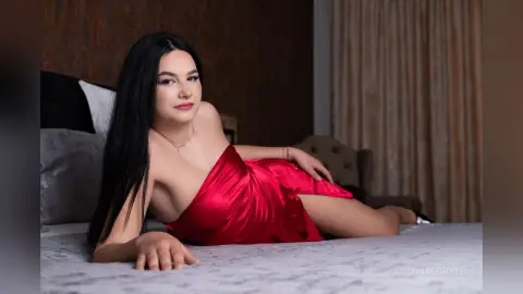 Young woman in a red satin dress relaxing on a bed in a moody, dimly lit bedroom setting.