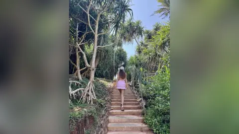 Young woman in a pink skirt walking up stone stairs through a lush tropical jungle forest.