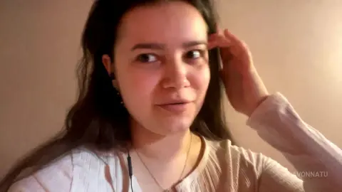 Young woman with long dark hair wearing a white ribbed sweater in a warm, cozy indoor setting.