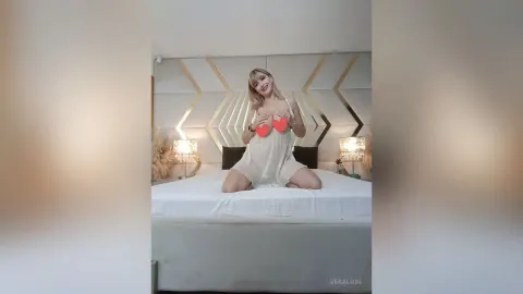 VeraLuxe — VeraLuxe sits on a white bed in a short white dress, holding red hearts over her bare breasts.