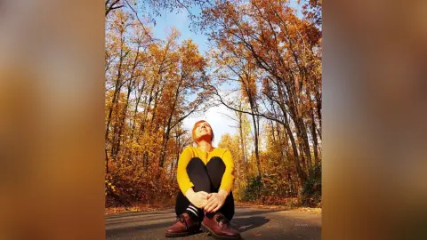 AryaSin sits on a road surrounded by autumn trees, wearing a yellow long-sleeve shirt, black leggings, and dark boots while looking up at the sky.
