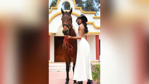 Elegant woman in a white flowing dress standing beside a brown horse in a sunny Mediterranean courtyard.
