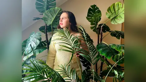 DariaCalm poses behind large green monstera leaves, wearing a light yellow mesh or net-style top or dress.