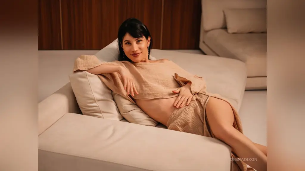 Young woman in white lace lingerie posing on a beige sofa in a warm, cozy indoor setting.