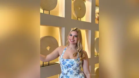 Smiling blonde woman in a blue floral sundress standing in a modern minimalist interior with warm lighting.