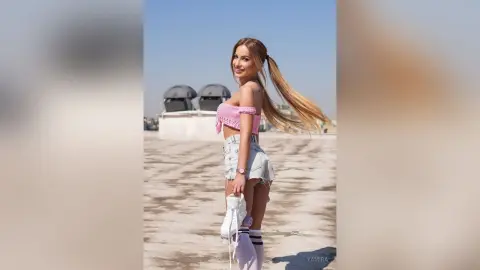 Smiling young woman in a pink crochet crop top and denim shorts posing on a sunny rooftop.