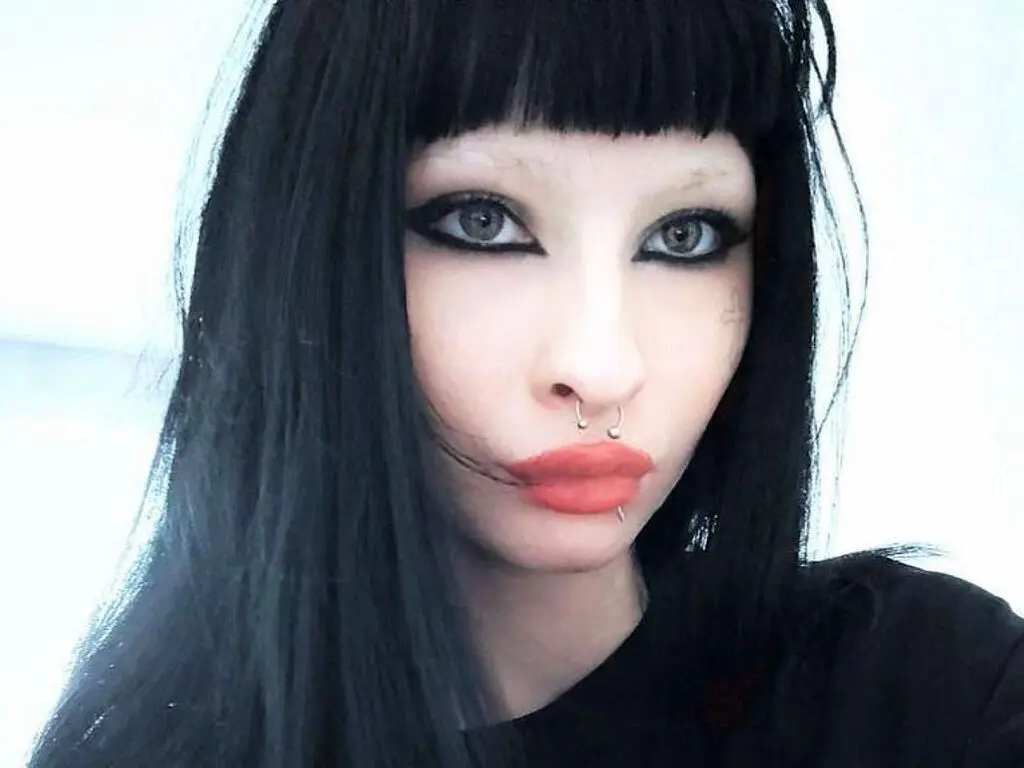 Goth woman with long black hair, heavy eyeliner, and lip piercings posing in a moody close-up portrait.