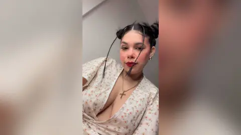 Smiling woman with space buns in a white floral wrap dress posing for a selfie in a bright room.
