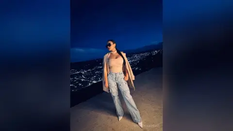Stylish woman in light blue jeans and beige top posing at night overlooking a glowing city skyline.
