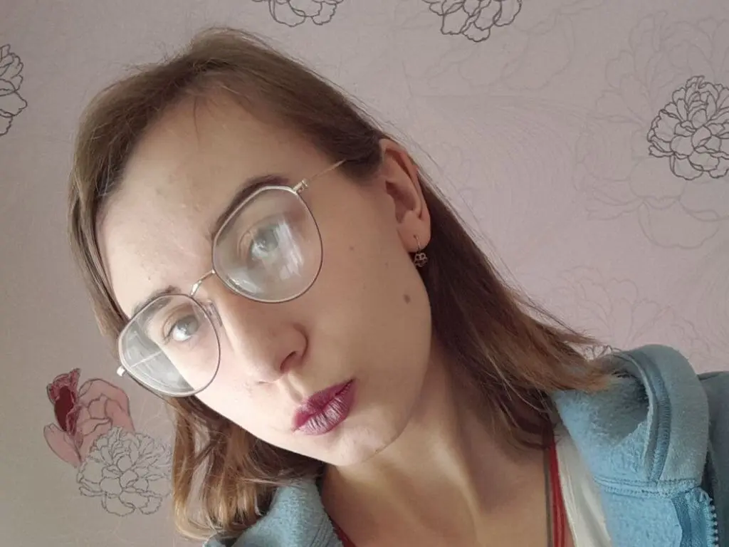 Young woman with glasses and light hair wearing a blue hoodie against a floral patterned background.