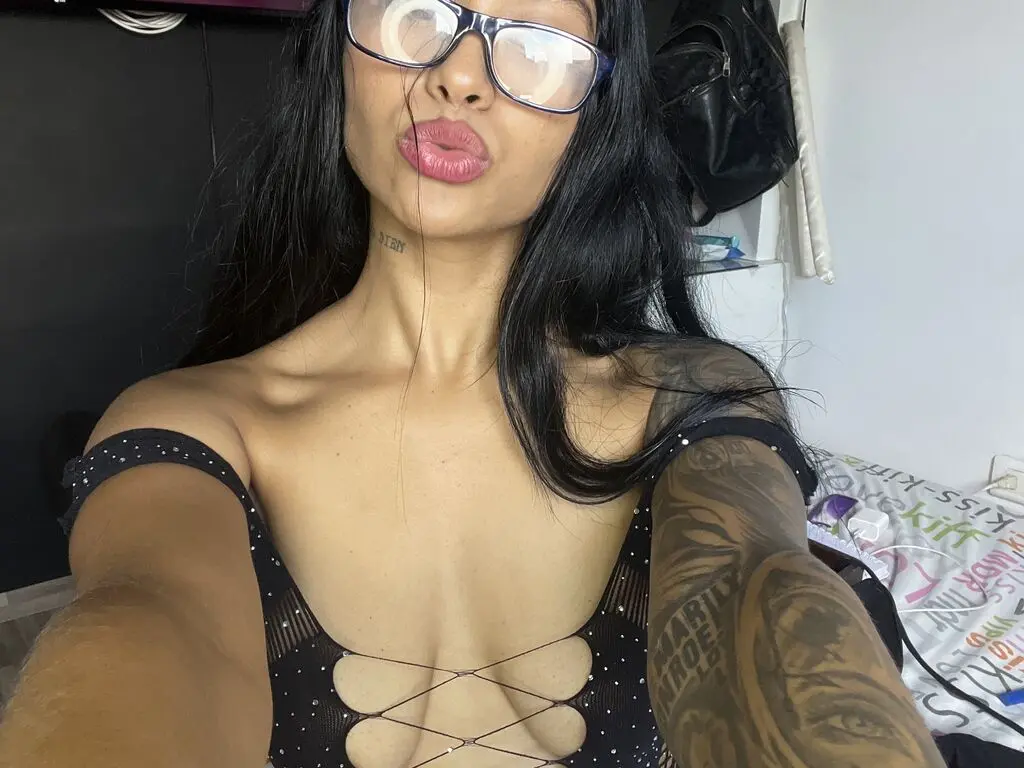 Young woman with long dark hair and glasses wearing a black strappy top in a casual indoor setting.