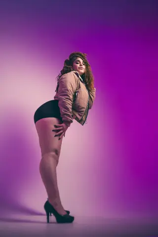 Confident woman in a tan jacket and black shorts posing against a vibrant purple studio background.