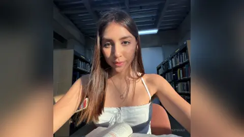 KateGale with long brown hair wearing a white camisole, sitting in a library setting with bookshelves behind her.