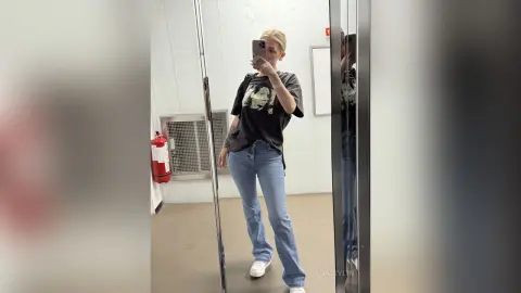 Blonde woman in a black graphic t-shirt and blue flared jeans taking a mirror selfie in a minimalist fitting room.