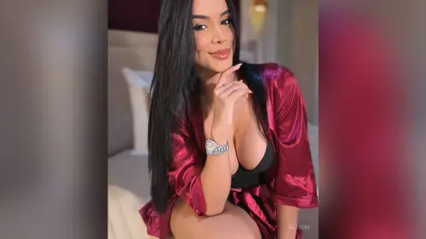 Alison poses in a red satin robe and black bra, resting her chin on her hand while looking at the camera.