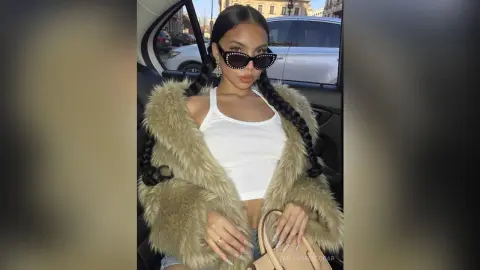 Stylish woman in a white tank top and tan faux fur coat wearing sunglasses sitting in a car.